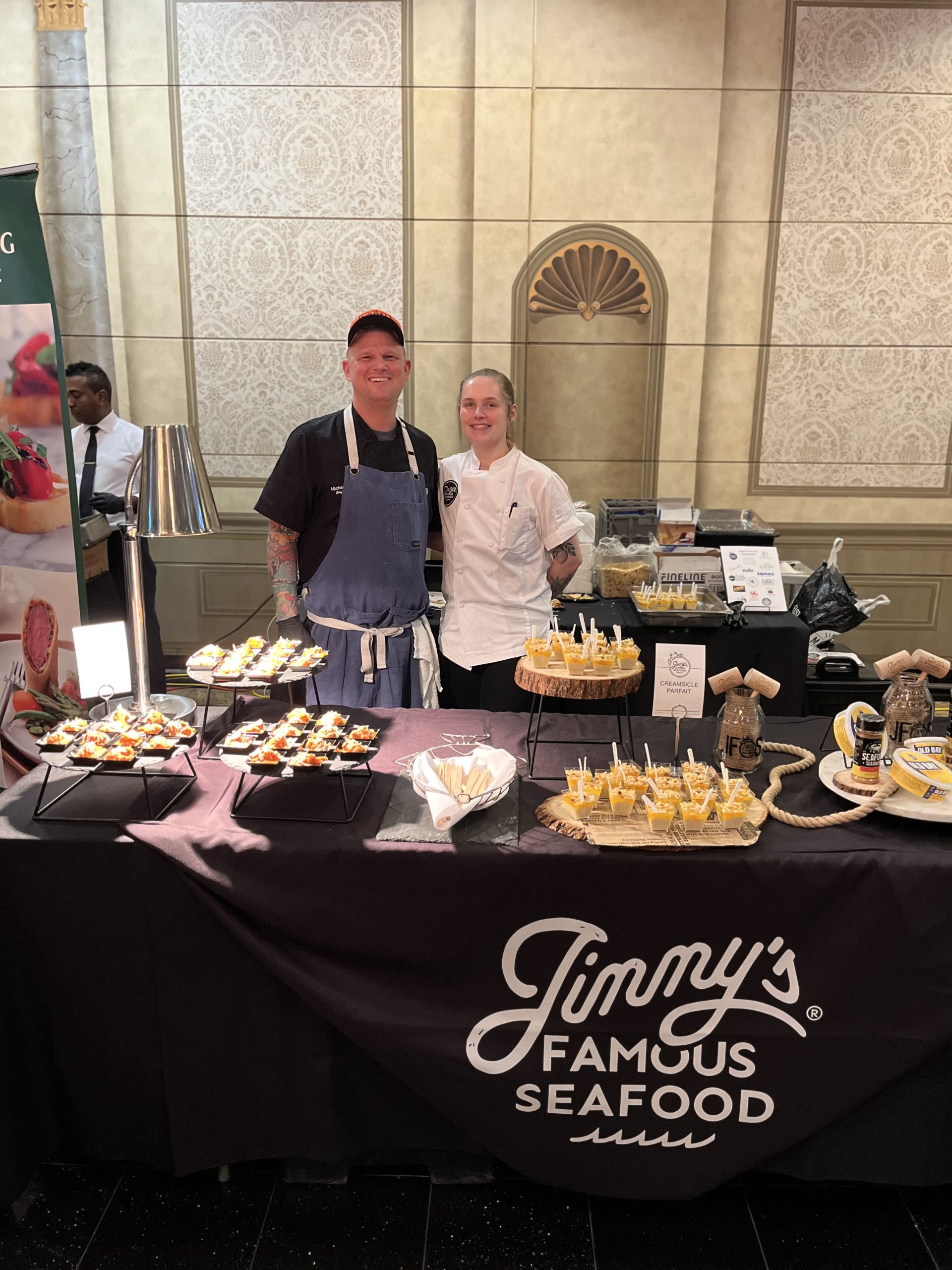 File_054 - Weekend Backpacks Baltimore Two chefs posed behind a table of their food samples with a sign saying "Jimmy's Famous Seafood."