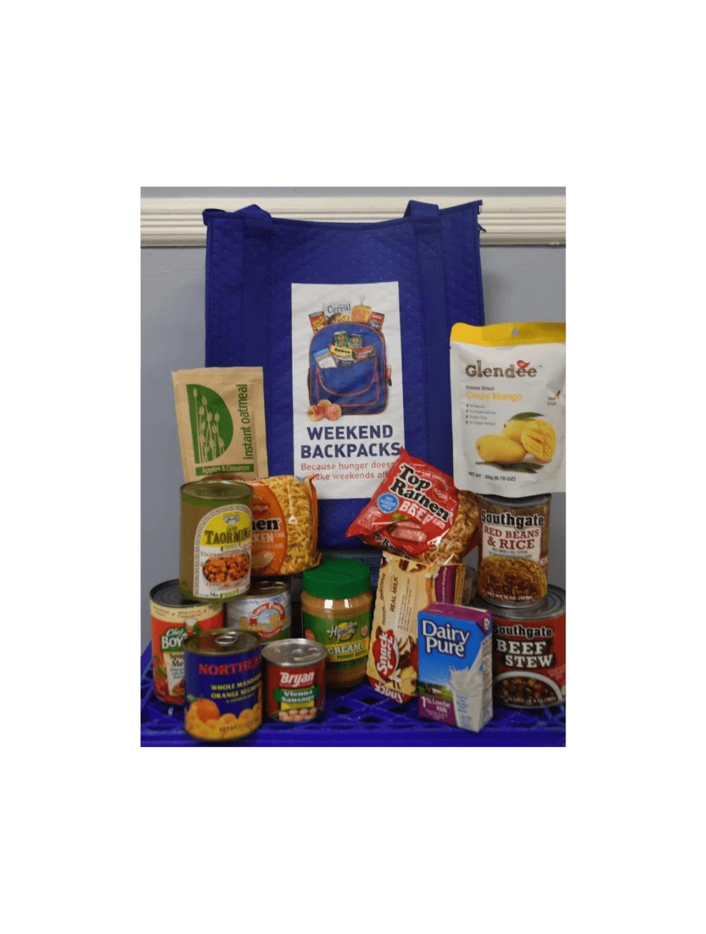Various non-perishable foods including peanut butter, ramen noodles, milk, oatmeal, a can of fruit, and prepared meals in front of a tote bag with the Weekend Backpacks logo.